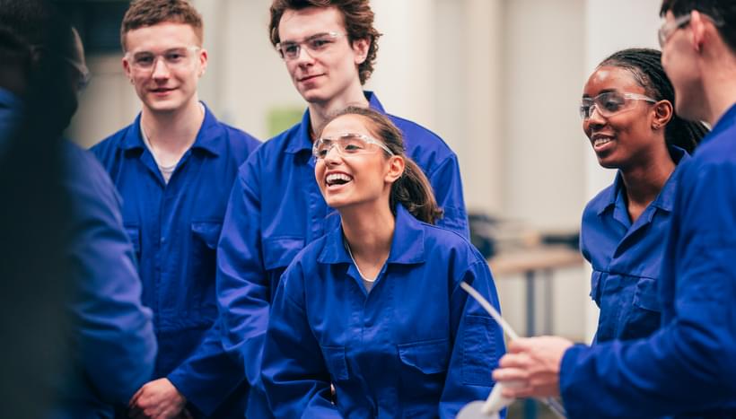 A group of young people in blue boiler suits and protective glasses are smiling and laughing