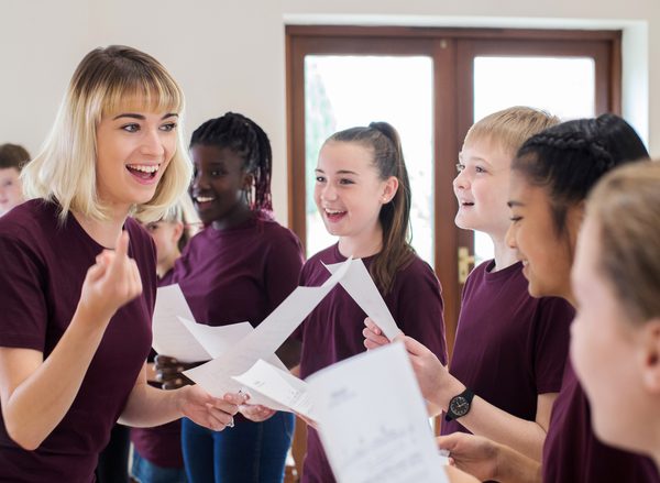 A group of teenage students wearing maroon t-shirts smiling and responding to a teacher during a rehearsal