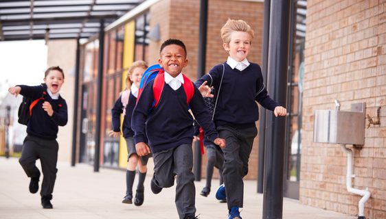 Happy primary school kids, wearing school uniforms and backpacks, running on a walkway outside their school building