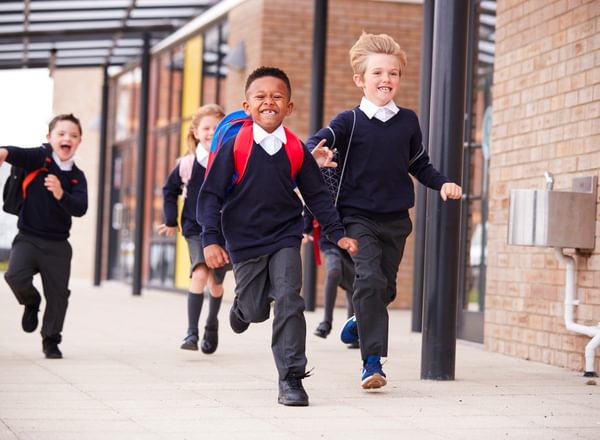 Happy primary school kids, wearing school uniforms and backpacks, running on a walkway outside their school building