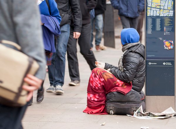 Female beggar in London. Sitting on a bag, with a blanket round her legs and a blue hood and brown coat. A newspaper and litter is on the path as she rests with a hand out against a map as people walk past