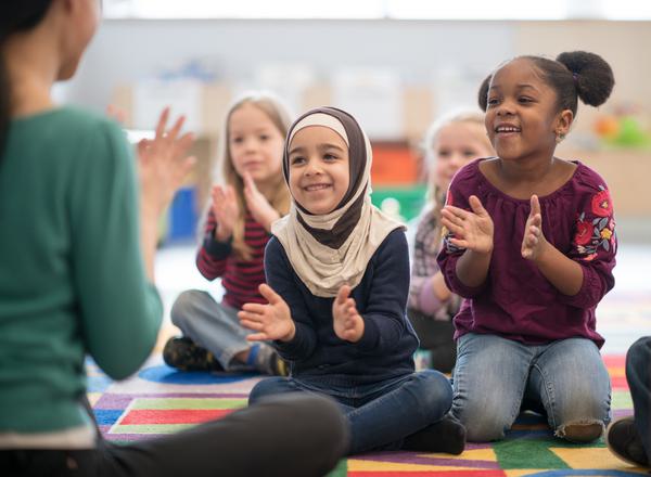 A diverse group of children sit in a colourful nursery setting, clapping along with the childminder