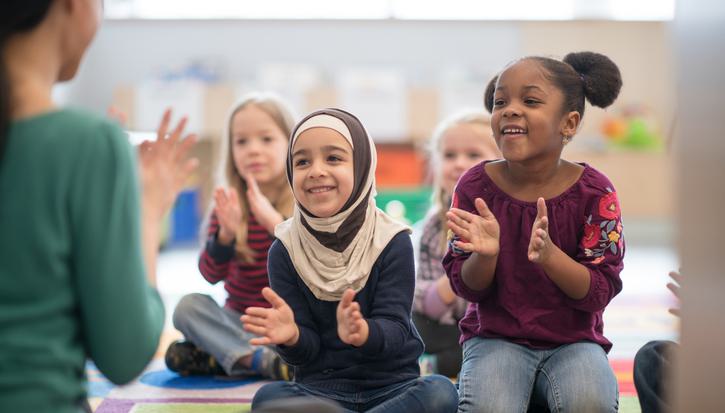 A diverse group of children sit in a colourful nursery setting, clapping along with the childminder