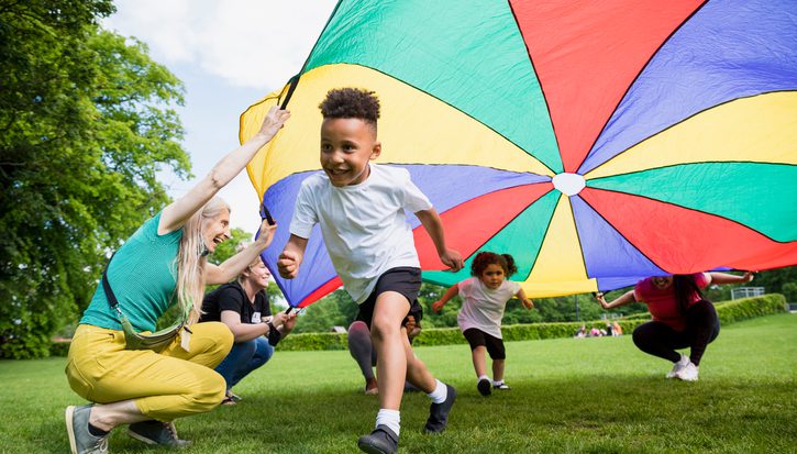 Two young children run playing underneath a rainbow canopy in a park or garden