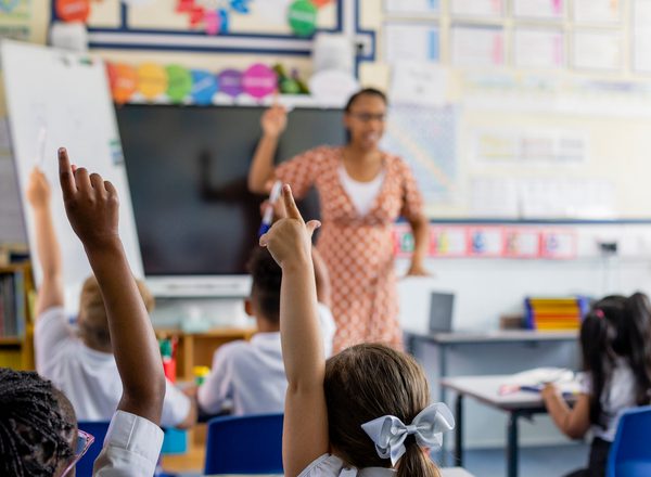 Primary school students sitting in a classroom being taught by a teacher in the North East of England. The students are answering questions with their arms in the air, the focus being on their arms.