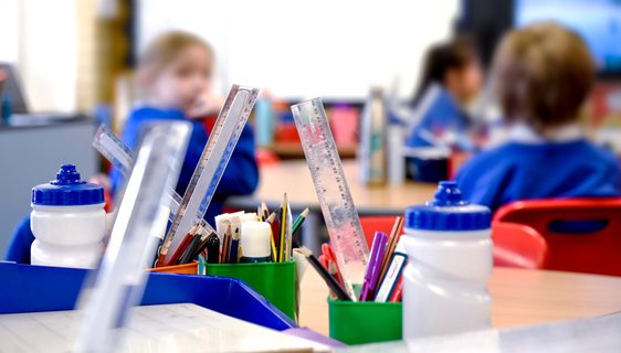 A primary school classroom. In the foreground are pots of pencils and rulers and water bottles. Children in blue school uniform are blurred in the background