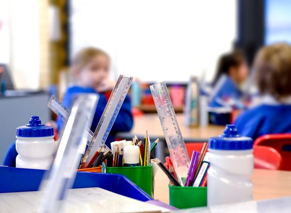 A primary school classroom. In the foreground are pots of pencils and rulers and water bottles. Children in blue school uniform are blurred in the background
