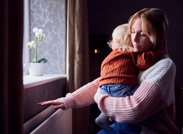 Mother With Son Trying To Keep Warm By Radiator At Home During Cost Of Living Energy Crisis