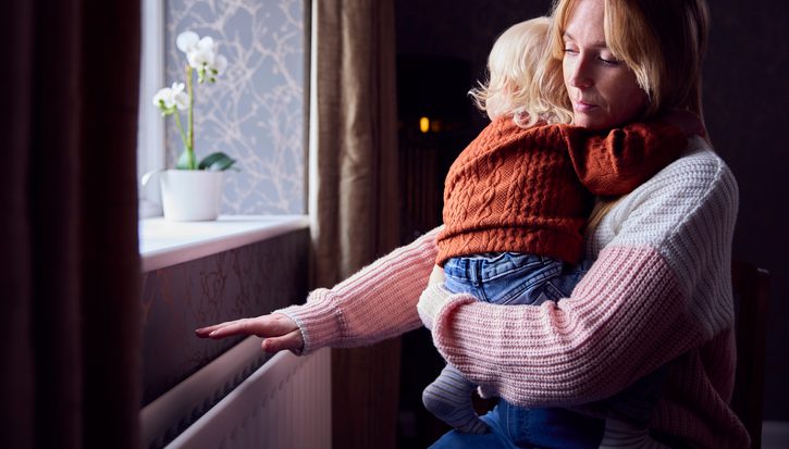 Mother With Son Trying To Keep Warm By Radiator At Home During Cost Of Living Energy Crisis