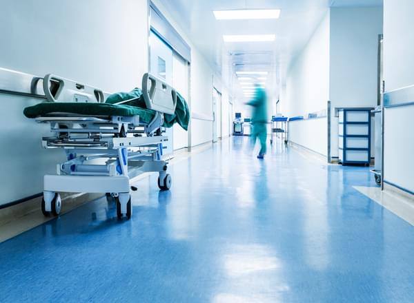A photo of a hospital corridor. A bed on wheel sits waiting while a person in scrubs runs away blurred in the distance