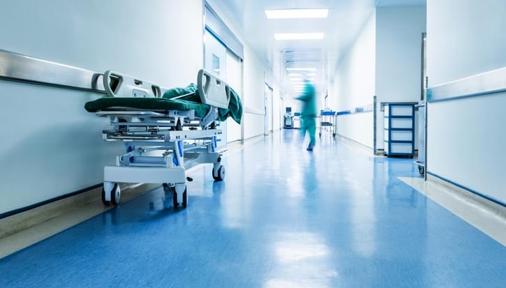 A photo of a hospital corridor. A bed on wheel sits waiting while a person in scrubs runs away blurred in the distance
