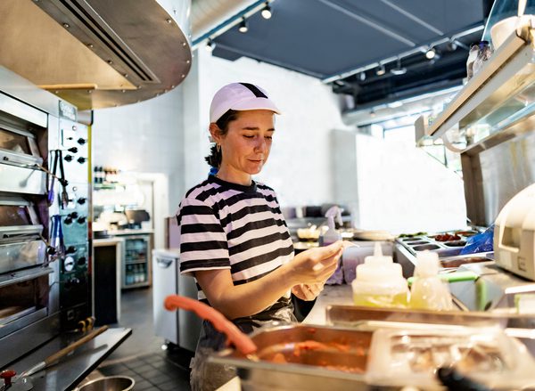 A woman in a white cap and black and white stripe t-shirt works in a restaurant kitchen