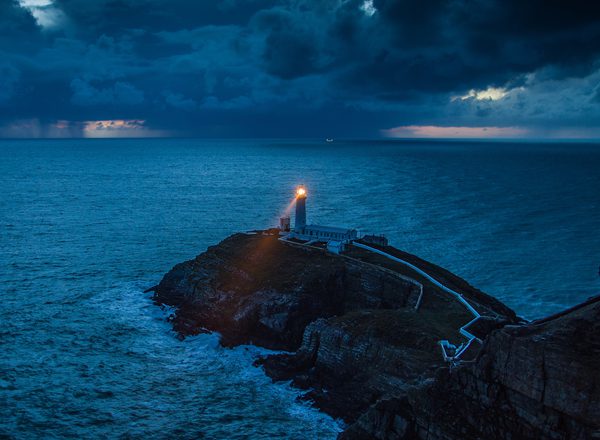 A lighthouse shines a light into a dark seascape at the edge of a cliff on the coast of Wales