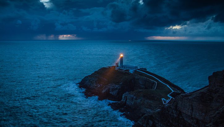 A lighthouse shines a light into a dark seascape at the edge of a cliff on the coast of Wales