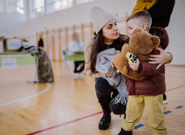 A woman with long dark hair wearing a woolly hat puts her arm around her small son who is holding a teddybear. They're in a community centre in front of boxes of charitable donations