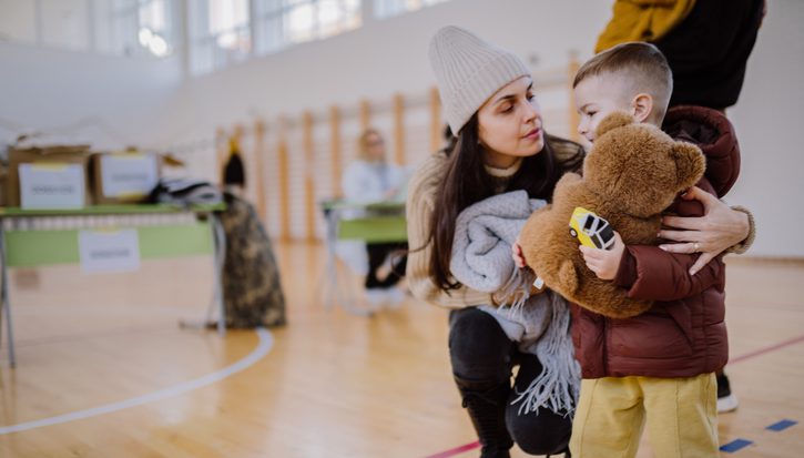 A woman with long dark hair wearing a woolly hat puts her arm around her small son who is holding a teddybear. They're in a community centre in front of boxes of charitable donations