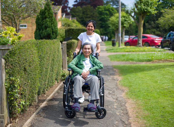 A female carer pushes an older female wheelchair user along a path in a residential area on a sunny day