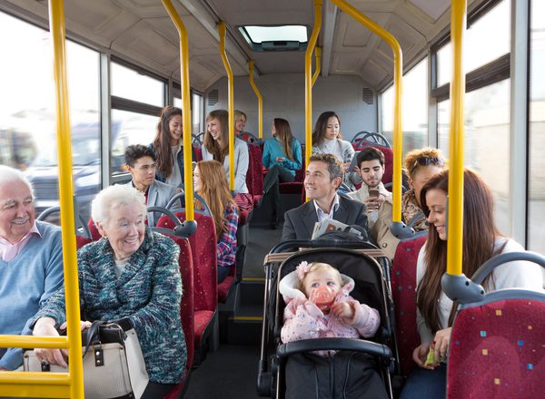 A busy bus full of people of all ages. An older couple smile at a younger woman with a baby in a pushchair