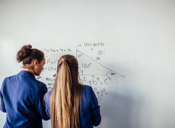 Two teenage girls can be seen from behind wearing blue school blazers working out a maths equation on a whiteboard
