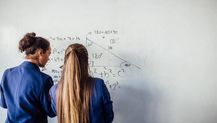Two teenage girls can be seen from behind wearing blue school blazers working out a maths equation on a whiteboard