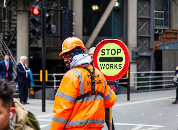 A Black man in a bright orange high vis jacket and hard hat stands in the middle of a road holding a sign that says 'STOP - WORKS'