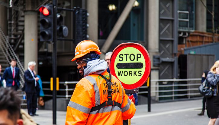 A Black man in a bright orange high vis jacket and hard hat stands in the middle of a road holding a sign that says 'STOP - WORKS'