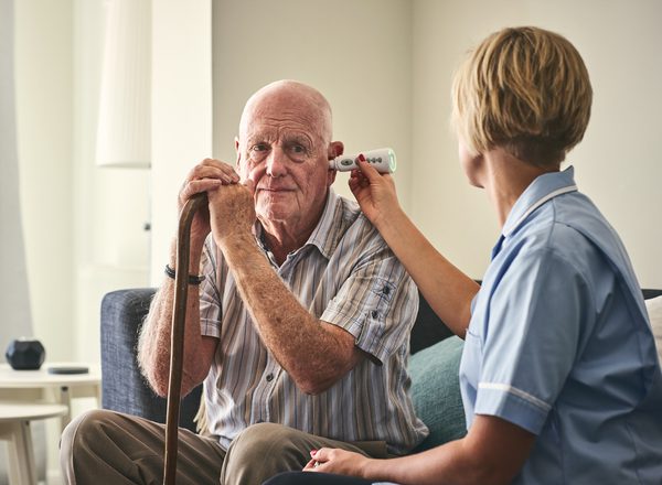 An older man with a walking stick is sitting on a sofa having his temperature taken with a thermometer in his ear by a female nurse in uniform