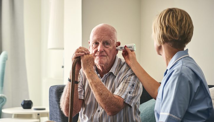 An older man with a walking stick is sitting on a sofa having his temperature taken with a thermometer in his ear by a female nurse in uniform