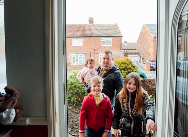 A young boy and girl entering the front door of a house, smiling. Their father carries a baby behind them