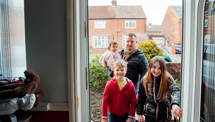 A young boy and girl entering the front door of a house, smiling. Their father carries a baby behind them