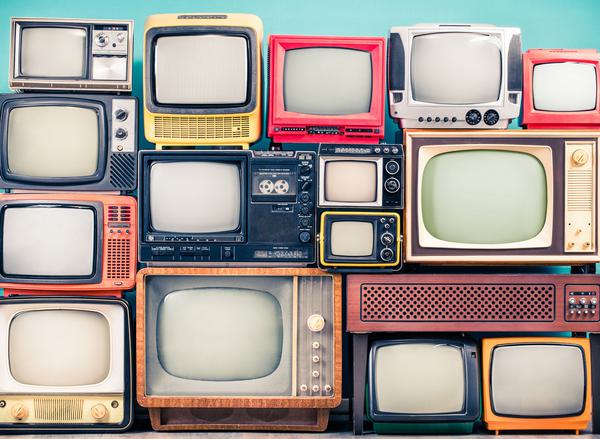 A stack of differently-shaped and colourful old-fashioned TVs from different eras in front of a blue background