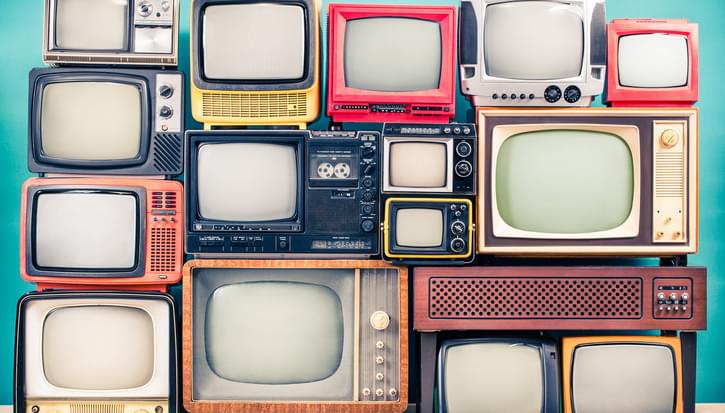 A stack of differently-shaped and colourful old-fashioned TVs from different eras in front of a blue background