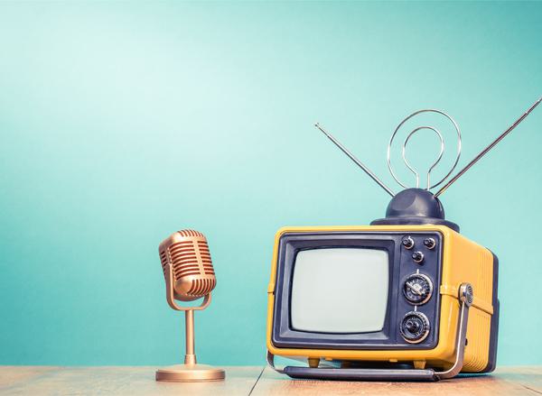 An old fashioned yellow TV and standing microphone sit on a desk in front of a turquoise background