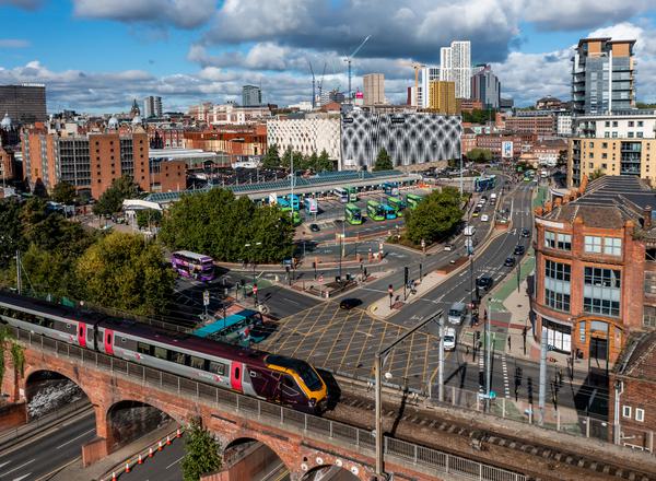 A high-up view of Leeds city centre: a bustling city with a train over a viaduct in the goreground and the city and lots of green buses behind