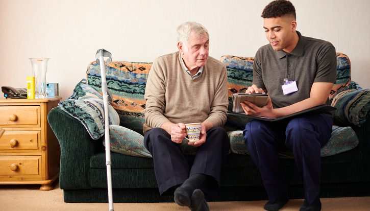 A young man speaks to an elderly man sat next to him on a sofa whilst pointing at a tablet computer